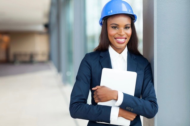 A woman in business attire holding papers and wearing a hard hat.